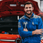 un meccanico in tuta blu e grigia sorridente a mezzo busto con braccia incrociate appoggiato al cofano di un'auto rossa con una chiave inglese in mano