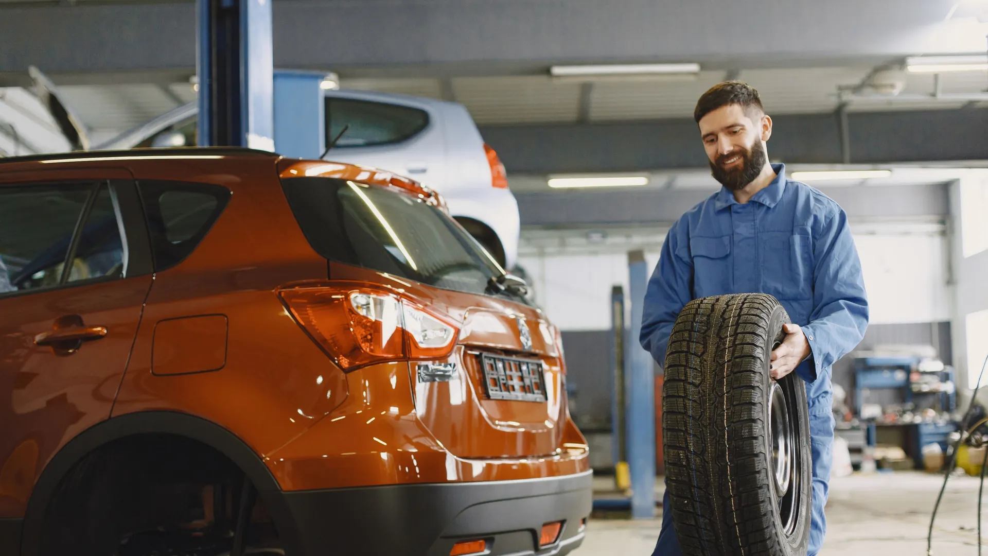Meccanico in tuta da lavoro che porta una gomma nuova accanto a un'auto in officina